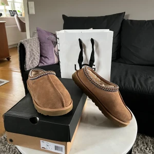 Bruna UGG Tasmans  - Säljer ett par bruna slip-in tofflor från UGG i mocka med mönstrad kant runt öppningen. Insidan är fodrad för extra komfort och sulan är robust och mönstrad. Perfekta för att hålla fötterna varma hemma eller utomhus. Skorna är knappt använda, köpta i januari på UGG Stockholm. Inget fel eller slitage på skorna. Skriv i dm för flera bilder eller förhandling av pris. Storleken är 44 EU. Kvitto, box, och påse finns även. 