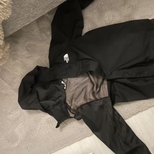 Svart vindjacka från The North Face - Säljer en svart vindjacka från The North Face med huva och dragkedja. Jackan har meshfoder på insidan och klassisk logga på bröstet. Perfekt för blåsiga dagar och enkel att matcha med det mesta.