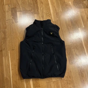 Svart väst från Lyle & Scott - Säljer en svart väst från Lyle & Scott med dragkedja framtill och två fickor med dragkedja. klassiska Lyle & Scott-loggan på bröstet. Till skillnad från många andra västar är kragen på västen lite högre så man kan vika ner eller ha den upp.