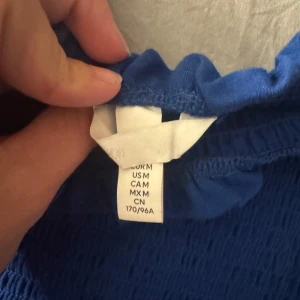 Blå smockad tubtopp med volang - Säljer en blå smockad tubtopp med volangkant upptill och nedtill. Toppen är elastisk och sitter tight mot kroppen, perfekt för sommardagar. Enkel att matcha med både kjol och jeans.
