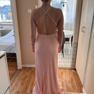 Ljusrosa glittrig långklänning med öppen rygg - Säljer en ljusrosa långklänning med glittrigt tyg och djup v-ringning. Klänningen har smala axelband som korsas i ryggen och en öppen rygg. Figurnära passform och lätt utsvängd nertill, perfekt för bal eller fest.