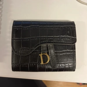 Svart korthållare med krokodilpräglat mönster - Säljer en svart plånbok  i skinn med krokodilpräglat mönster och guldfärgad D-detalj framtill. Plånboken har flera fack för kort och en praktisk knappstängning. Använt med lite slitage men i bra skick!