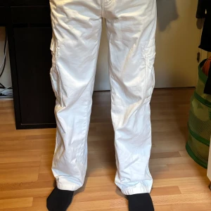 Vita cargopants med raka ben - Säljer ett par vita cargopants med raka ben och flera praktiska fickor på sidorna. Byxorna har normal passform och är tillverkade i bomull, perfekta för en avslappnad stil. Klassisk midja och diskreta sömdetaljer.
