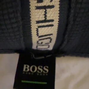 Svart hoodiejacka från BOSS - Svart hoodiejacka från BOSS med dragkedja framtill och vit logga på bröstet. Jackan har huva med snörning och vita detaljer längs dragkedjan och ärmarna. Perfekt för en avslappnad stil.