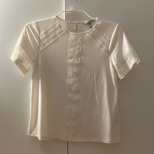 Vit topp med spetsdetaljer från H&M - Säljer en vit topp från H&M med fina spetsdetaljer längs ärmar och framtill. Toppen har korta ärmar och rund halsringning. Perfekt för en fräsch och stilren look.