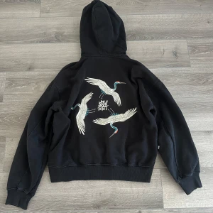 Svart hoodie Sweet Sktbs - Säljer en svart hoodie från Sweet Sktbs med broderade tranor och japansk text på ryggen. Tröjan har huva och klassisk känguruficka framtill. Perfekt för dig som gillar streetwear med unika detaljer.
