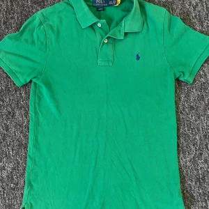 Grön pikétröja från Polo Ralph Lauren - Säljer en klassisk grön pikétröja från Polo Ralph Lauren i slim fit-modell. Tröjan har korta ärmar, krage och knappar framtill samt den ikoniska broderade loggan på bröstet. Perfekt för en stilren och avslappnad look.