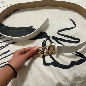 Vitt bälte med guldspänne - Säljer ett vitt bälte med ett unikt, dekorativt spänne i guld. Bältet har en stilren design och är tillverkat i ett slätt material. Perfekt accessoar för att lyfta din outfit.