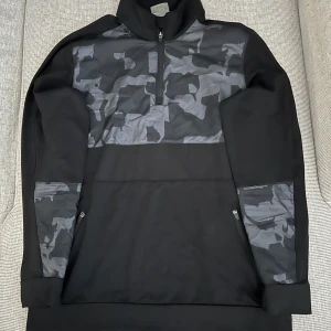 Svart tröja med camouflagedetaljer och halv dragkedja - Säljer en svart tröja med halv dragkedja och camouflagemönster över bröstet och på ärmarna. Insidan av kragen har en grön detalj. Perfekt för en sportig och avslappnad stil.