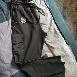 Svarta cargopants från OFF - Säljer ett par svarta cargopants från OFF med elastisk midja och dragsko. Byxorna har stora fickor på sidorna, dekorativa detaljer med dragkedja och nyckelring samt OFF-logga på ena benet. Perfekta för en streetwear-look.