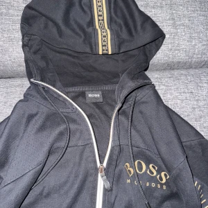Svart hoodiejacka från Hugo Boss - Snygg svart hoodiejacka från Hugo Boss med gulddetaljer och dragkedja framtill. Jackan har huva med snörning och tryckt logga på bröstet. Perfekt för en stilren och sportig look.