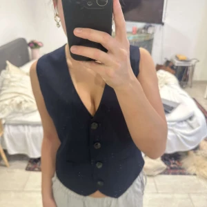 Svart väst med knappar - Säljer en svart väst med knäppning framtill och v-ringad hals. Västen har en klassisk passform och är utan ärmar, perfekt att styla över en skjorta eller t-shirt.