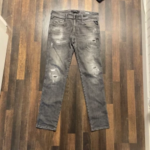 Grå Replay Anbass jeans med slitningar - Säljer ett par mörkgråa jeans från Replay, modell Anbass. Byxorna har en slim passform och är dekorerade med slitningar och detaljerade sömmar. Klassisk femficksmodell med Replay-logga på fickan.