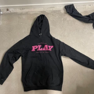 Svart hoodie från Playboy med rosa tryck - Säljer en svart hoodie från Playboy med stort rosa PLAY-tryck på bröstet och textdetaljer. Mjuk fleece på insidan och klassisk känguruficka framtill. Perfekt för en avslappnad och cool stil.