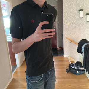 Svart pikétröja från Ralph Lauren - Klassisk svart pikétröja från Ralph Lauren med liten röd broderad logga på bröstet. Tröjan har krage och korta ärmar, perfekt till jeans eller shorts. Snygg och stilren design som passar till mycket.