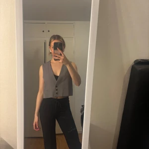 Grå väst med knappar - Snygg grå väst med v-ringning och knäppning framtill. Västen har en rak passform och är croppad, vilket ger en modern look. Perfekt att styla över en topp eller skjorta.