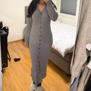 Grå ribbad långklänning med knappar - Säljer en grå långklänning i ribbat material med v-ringning och långa ärmar. Klänningen har knappar hela vägen framtill och en slits nedtill. Perfekt för dig som gillar en tight passform och stilren look. Går att använda som cardigan. Inte använd alls och är adoore liknande 