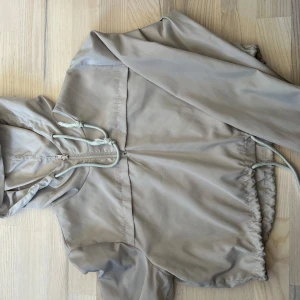 Beige vindjacka från PrettyLittleThing - Säljer en beige croppad vindjacka från PrettyLittleThing med huva, dragkedja och dragsnöre i midjan. Jackan har långa ärmar och en avslappnad passform, perfekt för blåsiga dagar.