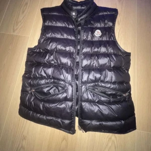 Svart dunväst från Moncler. !! Har Scan kod på!! - Säljer en svart dunväst från Moncler med hög krage och dragkedja framtill. Västen har två snedställda fickor med dragkedja och Moncler-logga på bröstet. Perfekt för lager-på-lager och stilren look. Lite komplicerat me dragkedjan men o liten sytt på ryggen men ingen som syns när man har på sig 