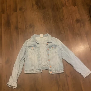 Ljusblå jeansjacka med knappar - Säljer en klassisk ljusblå jeansjacka med silverfärgade knappar och två bröstfickor. Jackan har en croppad passform och långa ärmar. Perfekt att slänga över en t-shirt för en avslappnad look.