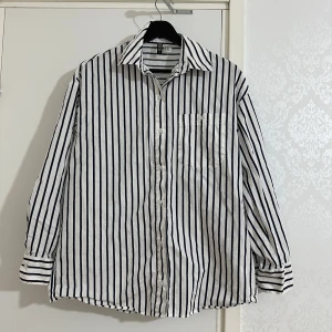 Vit och marinblå randig skjorta från H&M - Vit skjorta med marinblå vertikala ränder från H&M. Klassisk krage, knäppning framtill och bröstficka. Långärmad modell i bomull, perfekt för en stilren och avslappnad look.  storlel Xs till m