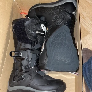 Svarta alpine stars cross stövlar - Säljer ett par svarta alpinestars Adventure kängor med högt skaft och spännen. Skorna har robust design, vadderad insida och är perfekta för utomhusaktiviteter. Tillverkade i syntetmaterial och har en platt sula för extra stabilitet.