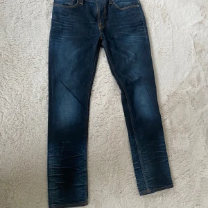 Mörkblå jeans från Nudie Jeans - Säljer ett par mörkblå jeans från Nudie Jeans med klassiska orange sömmar och snygga slitningar. Jeansen har rak passform och normal midja. Perfekta för dig som gillar stilrena och tidlösa jeans.