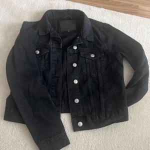 Svart jeansjacka med silverknappar - Säljer en klassisk svart jeansjacka med silverfärgade knappar och två bröstfickor. Jackan har en rak passform och krage, perfekt att slänga över en hoodie eller t-shirt. Enkel och tidlös design som funkar till det mesta.