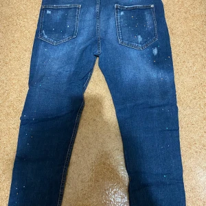 Mörkblå dsquared jeans med färgstänk - Säljer ett par mörkblå jeans med hål på framsidan samt små färgstänk över hela byxan. Klassisk femficksmodell med raka ben och normal passform. Perfekta för dig som gillar en lite mer edgy stil.