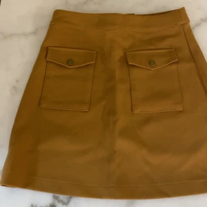 Beige kjol med fickor - Säljer en beige kjol med två stora fickor framtill och knappdetaljer. Kjolen har en A-linjeformad siluett och är perfekt för dig som gillar en enkel men snygg stil. Passar till många olika outfits!