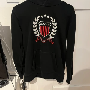 Svart hoodie från GANT med broderad logga - Svart hoodie från GANT med stor broderad logga och emblem i vitt och rött på bröstet. Tröjan har huva och magficka, samt ribbade muddar vid ärmslut och nederkant. Perfekt för en avslappnad och stilren look.