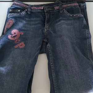 Jeans med rosa detaljer och paljetter - Säljer ett par blå jeans med rosa paljettbroderier och dekorativa detaljer på fickor och bälte. Byxorna har låg midja och bootcut-modell, samt unika rosa inslag som gör dem extra speciella. Dem är xs och har några slitage men inget som märks, dem är sprättade längst ner för att bli längre, jag är 165 och är lite korta på mig. Säljer för dem är lite små i midjan men kan fortfarande ha dem fast jag är ungefär storlek s.