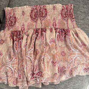 Rosa mönstrad kortkjol med volang - Superfin kortkjol i rosa och beige toner med paisleyinspirerat mönster. Kjolen har en smockad midja.🩷pris kan diskuteras