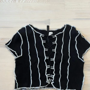 Svart ribbad croppad topp med snörning från H&M Divided - Säljer en svart croppad topp från H&M Divided med vita kontrastsömmar och snörning framtill. Toppen är ribbad och har korta ärmar, perfekt för en trendig look.