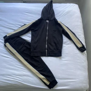 Svarta mjukisbyxor med vita sidor från Moncler - Finns även kvitto 🧾.Svarta mjukisbyxor från Moncler med vita ränder längs sidorna. Byxorna har resår och snörning i midjan, muddar vid bensluten och två fickor fram. Moncler-logga broderad på vänster ben.