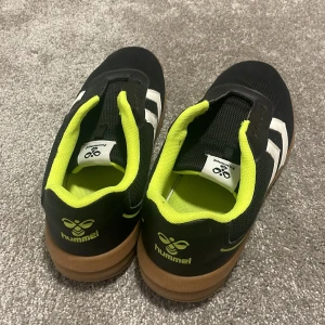 Svarta sneakers från Hummel med vita detaljer - Säljer ett par svarta Hummel sneakers med vita pilar på sidorna och brun sula. Insidan är neongul för en cool kontrast. Perfekta för dig som gillar sportig stil och vill sticka ut lite extra.