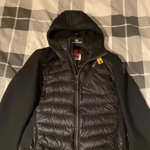 Svart dunväst med huva från Parajumpers - Snygg svart dunväst från Parajumpers med huva och dragkedja framtill. Västen har quiltad framsida och detaljer i gult vid halsen. Perfekt för lager-på-lager och har en sportig look.