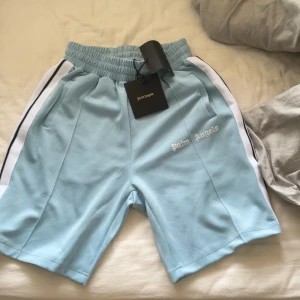 Ljusblå shorts från Palm Angels - Snygga ljusblå shorts från Palm Angels med vit logga broderad på ena benet. Shortsen har elastisk midja, vita och svarta ränder längs sidorna samt fickor på båda sidor. Perfekta för en avslappnad stil. Helt ny 
