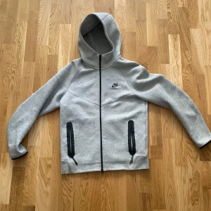 Grå Nike Tech Fleece - Snygg grå Nike Tech Fleece med svart dragkedja och svarta detaljer vid fickorna. Jackan har huva och diskret Nike-logga på bröstet. Perfekt för en sportig och avslappnad stil.
