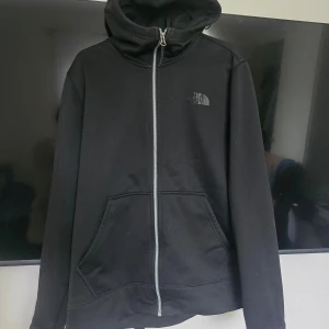 Svart hoodiejacka från The North Face - Svart hoodiejacka från The North Face med dragkedja och två stora fickor framtill. Jackan har huva och diskret logga på bröstet. Perfekt för en avslappnad stil och enkel att matcha med det mesta. Använd enstaka gång . Super skönt material  . Hundhår kan förekomma då hund finns i hem.  Sonen har storlek S-M i herrstorlek och ca 176 i ungdom så skulle säga den är inom de