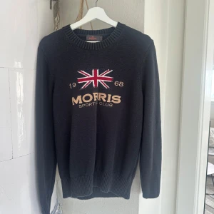 Mörkblå stickad tröja från Morris - Säljer en mörkblå stickad tröja från Morris med broderad Union Jack-flagga och texten 'Morris Sport Club 1968' på bröstet. Tröjan har rund halsringning och ribbade muddar vid ärmslut och nederkant.
