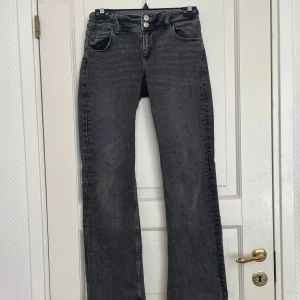 Svarta bootcut jeans med knappar - Säljer ett par svarta bootcut jeans med snygga detaljer som dubbla knappar i midjan och fickor med lock och knapp bak. Jeansen har en klassisk femficksdesign och är perfekta för dig som gillar en tidlös stil.