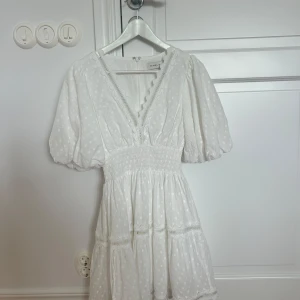 Vit kortklänning med puffärm från By Malina - Superfin vit kortklänning från By Malina med puffärmar, v-ringning och smock i midjan. Klänningen har diskret prickigt mönster, spetsdetaljer och volang nertill. Perfekt för student och konfirmation! Använd en gång 