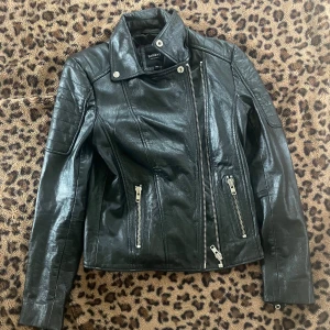 Svart skinnjacka från Barneys Originals - Snygg svart skinnjacka från Barneys Originals med klassisk bikerstil. Jackan har sned dragkedja, två dragkedjefickor framtill och markerad krage med tryckknappar. Perfekt för en cool och tidlös look.