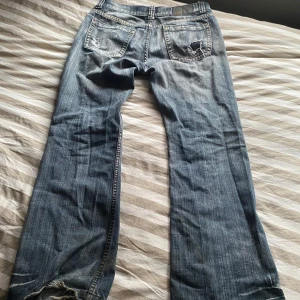 Feta Smog jeans - Ett par klassiska blå jeansbyxor från Smog med slitna detaljer på bakfickorna. Byxorna har normal passform och raka ben, perfekt för en avslappnad stil. Tydliga slitningar ger en cool, använd look. Stängs med knapp och dragkedja.