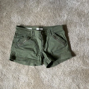 Olivgröna shorts med dragkedjedetaljer från Three Sixty Five - Snygga olivgröna shorts från Three Sixty Five med två snedställda fickor framtill som har dragkedjor. Klassisk knapp och gylf, bälteshällor och bakfickor. Perfekta för varma dagar och enkel att matcha med olika toppar🌸 