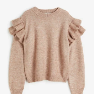 Beige stickad tröja med volang från H&M - Säljer en beige stickad tröja från H&M med långa ärmar och volangdetaljer på axlarna. Tröjan har rund halsringning och ribbade muddar vid ärmslut och nederkant. Mjukt och varmt material. Passar mig som har xs/s