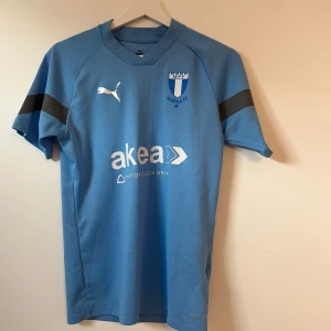 Malmö FF fotbollströja från Puma - Ljusblå Malmö FF matchtröja från Puma med klubbmärke på bröstet och svart detalj på ärmarna. Kortärmad modell med tryckt sponsorlogga på framsidan. Perfekt för fotbollsfans!