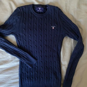 Mörkblå kabelstickad tröja från Gant - Säljer en klassisk mörkblå kabelstickad tröja från Gant med rund halsringning och broderad logga på bröstet. Tröjan har långa ärmar och ribbade muddar vid ärmslut och nederkant. Perfekt för en stilren och tidlös look.