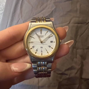 Seiko herrklocka i silver och guld - Klassisk Seiko herrklocka med tvåfärgad länk i silver och guld. Vit urtavla med romerska siffror, datumvisning och guldfärgad ring runt boetten. Armbandet är i metall med inslag av guldtonade detaljer. Nypris 2250kr. 